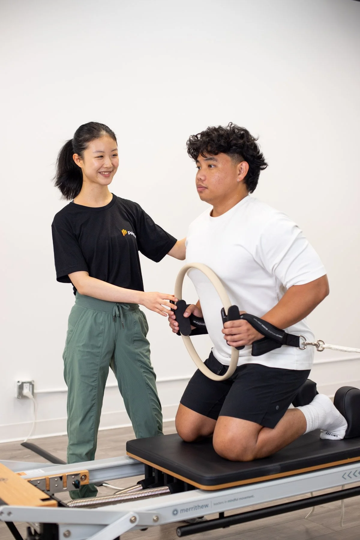 A woman providing physical therapy or injury treatment to a person lying on a table, wearing a black t-shirt with a logo, in a clinical setting.