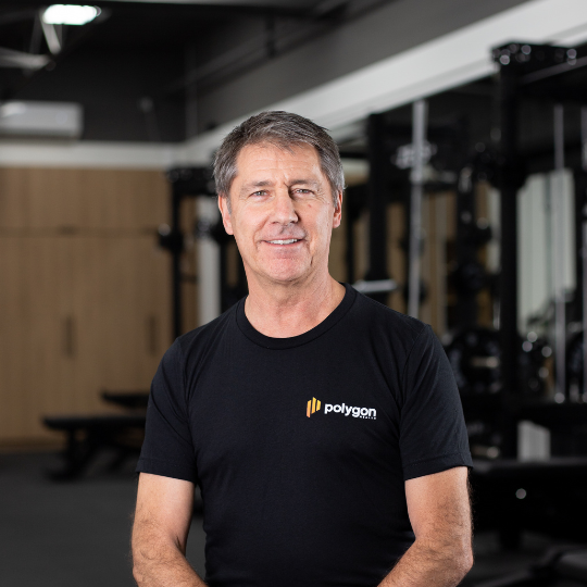 Middle-aged man standing in a gym, wearing a black t-shirt with 'polygon' logo, smiling.