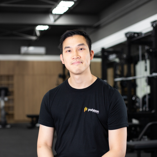 A young man standing in a gym, wearing a black t-shirt with a logo that says 'polywork'. He has short black hair and is smiling slightly.