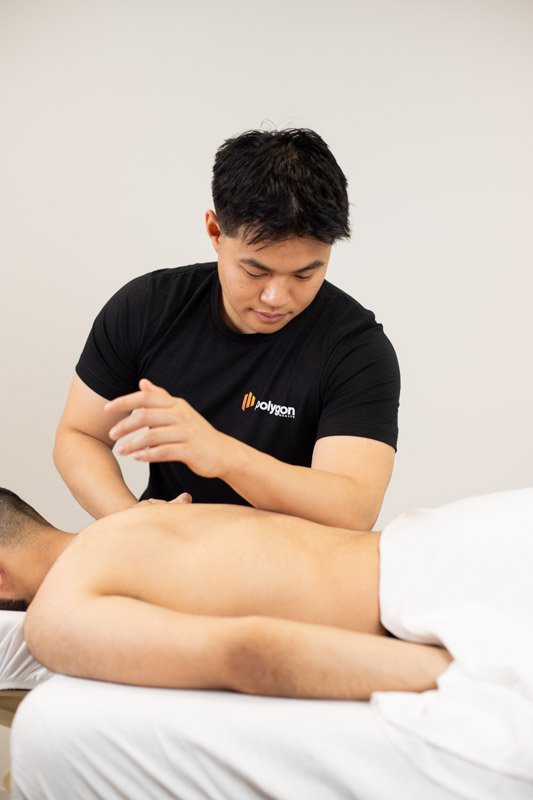 A woman providing physical therapy or injury treatment to a person lying on a table, wearing a black t-shirt with a logo, in a clinical setting.