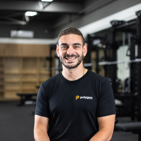 A smiling man in a black Polygon t-shirt standing in a gym with workout equipment in the background.