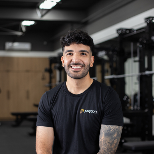 A smiling man with dark curly hair and a beard, wearing a black t-shirt with a Polygon logo, in a gym setting with workout equipment in the background.