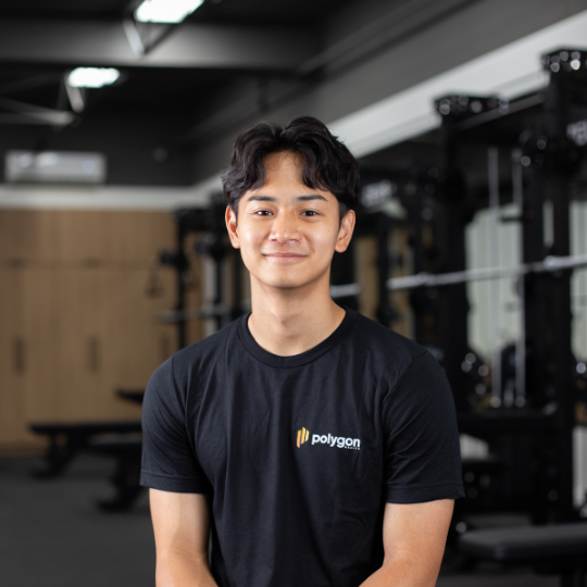 A young man with dark hair and a light smile standing in a modern gym, wearing a black t-shirt with the word 'Polygon' logo.