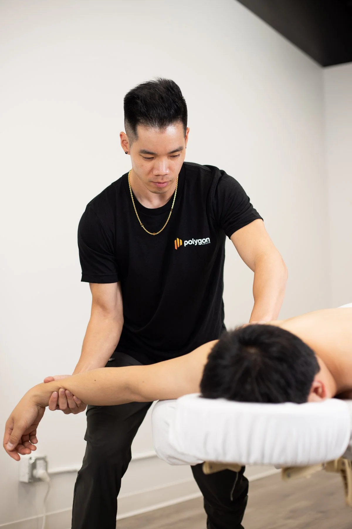A woman providing physical therapy or injury treatment to a person lying on a table, wearing a black t-shirt with a logo, in a clinical setting.