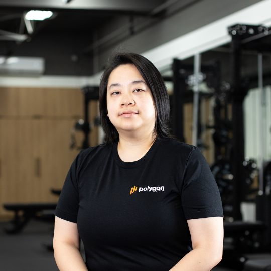 A woman with medium-length dark hair in a gym, wearing a black Polygon t-shirt, standing with arms crossed.