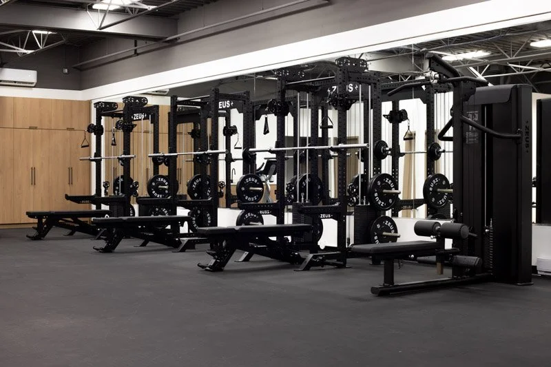 Empty gym with black weightlifting equipment and benches, light wood cabinets on the wall, and a gray floor.