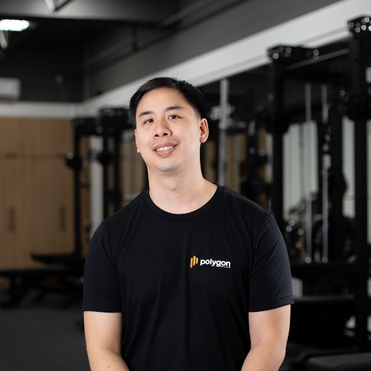 A young man with black hair smiling at the camera, wearing a black Polygon T-shirt, in a gym with fitness equipment in the background.