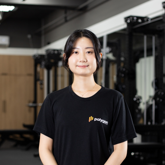 A young woman standing in a gym, wearing a black Polygon t-shirt, with gym equipment and mirrors in the background.