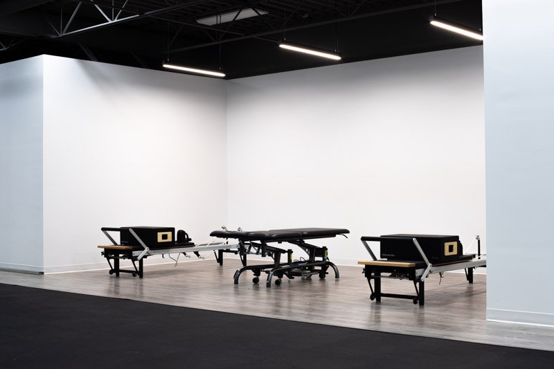 An art installation featuring three black benches with speakers and a massage table in an empty gallery with white walls and a black ceiling.