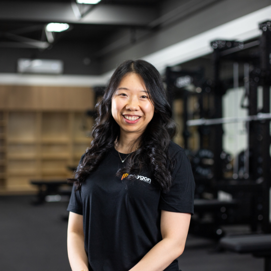 A smiling woman with long dark hair standing in a gym, wearing a black t-shirt.