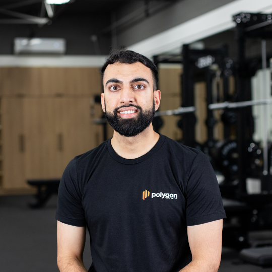 A man with dark hair and a beard smiling, wearing a black t-shirt that says 'polygon' on it, standing in a gym with exercise equipment in the background.