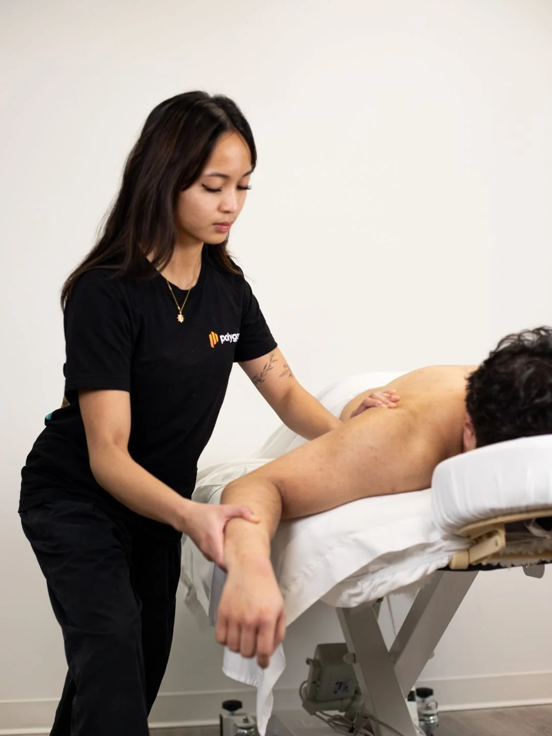 A woman providing physical therapy or injury treatment to a person lying on a table, wearing a black t-shirt with a logo, in a clinical setting.