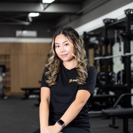 A young woman with wavy hair wearing a black t-shirt and smartwatch, sitting in a gym with workout equipment in the background.