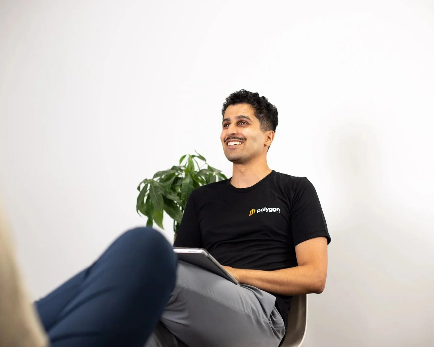 A smiling man sitting on a chair, holding a tablet, with a plant behind him, against a white wall.