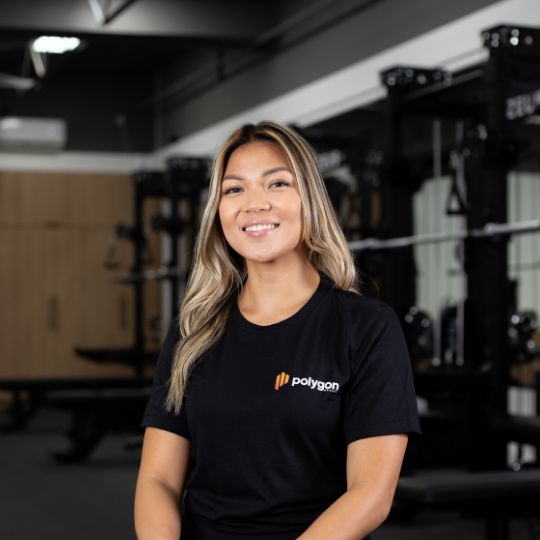 A woman with long blonde hair smiling in a gym, wearing a black polygon t-shirt.