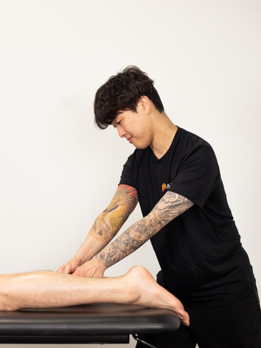 A woman providing physical therapy or injury treatment to a person lying on a table, wearing a black t-shirt with a logo, in a clinical setting.