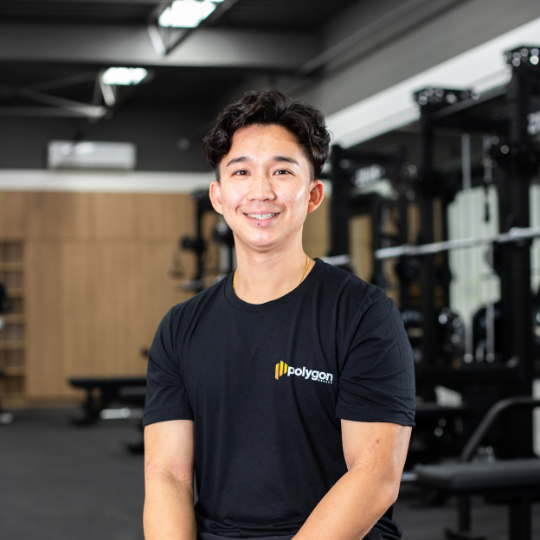 Young man smiling at the camera inside a gym, wearing a black T-shirt with a logo that says "Polygon".