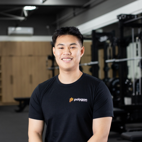 A young man smiling and standing in a gym, wearing a black Polygon t-shirt.