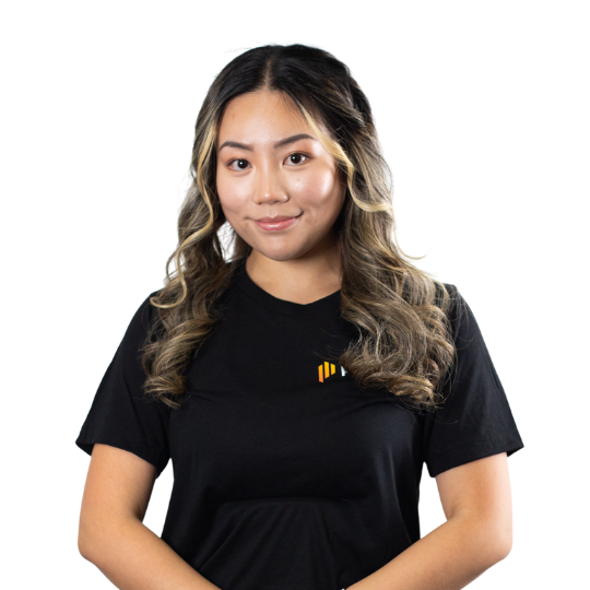 A young woman with long wavy hair wearing a black T-shirt with a small colorful logo, standing against a plain white background.