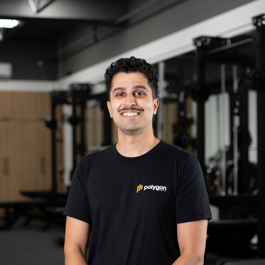 A smiling man with curly dark hair and a mustache wearing a black polygon t-shirt, standing in a gym with exercise equipment in the background.