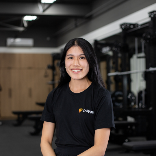 A young woman with long black hair smiling in a gym, wearing a black t-shirt with a logo that says 'polygap'.