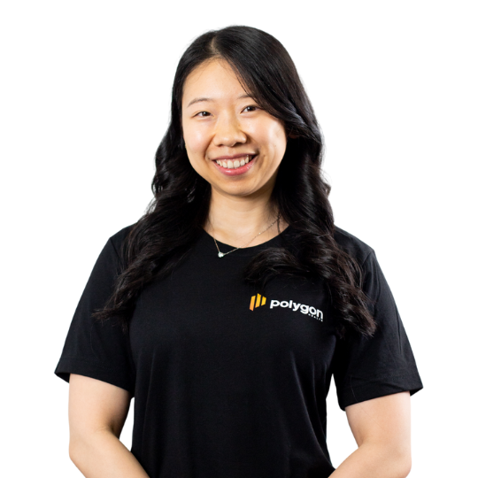 Young woman with long dark hair, smiling, wearing a black Polygon t-shirt and a necklace, standing against a plain background.