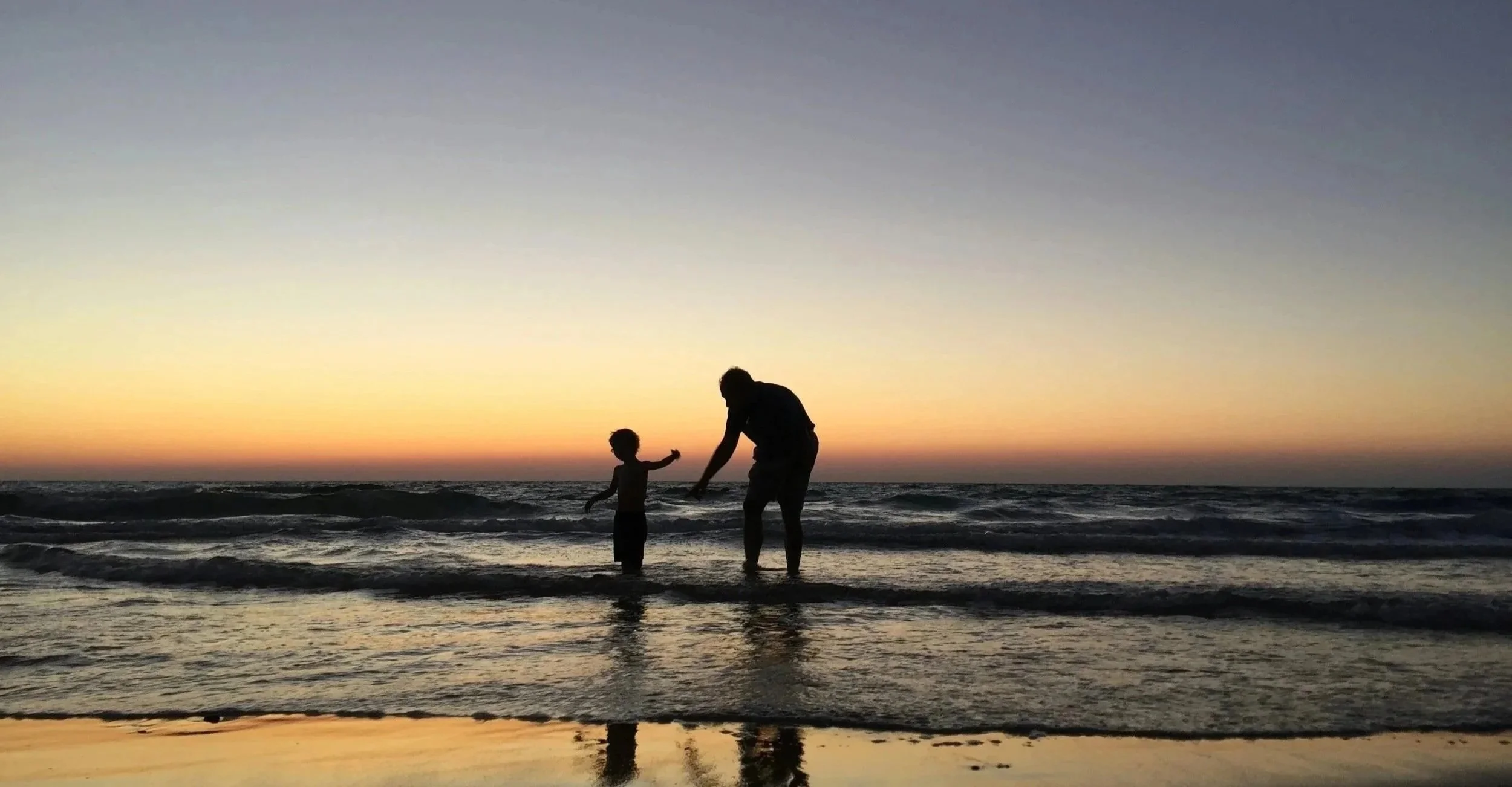 Silhouettes of an adult and a child playing with each other at the beach during sunset, with the ocean and colorful sky in the background.
