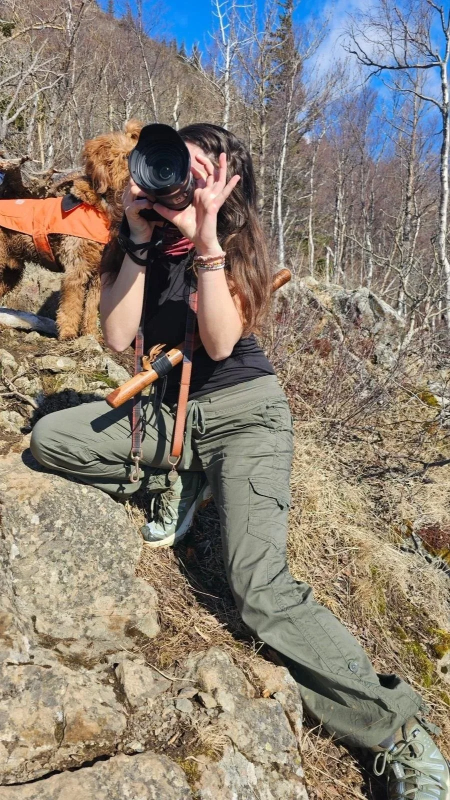 A woman with long brown hair holding a camera while kneeling on rocky ground in a forest with leafless trees, wearing green cargo pants, a black shirt, and sneakers, with a small brown dog wearing an orange vest beside her.