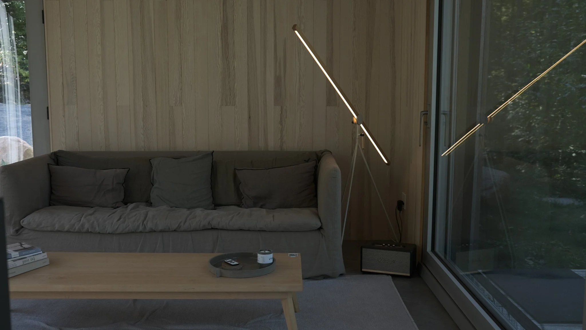 Living room with beige couch, cushions, wooden wall, tripod floor lamp, coffee table with items, large window with view of trees, and a Marshall speaker.