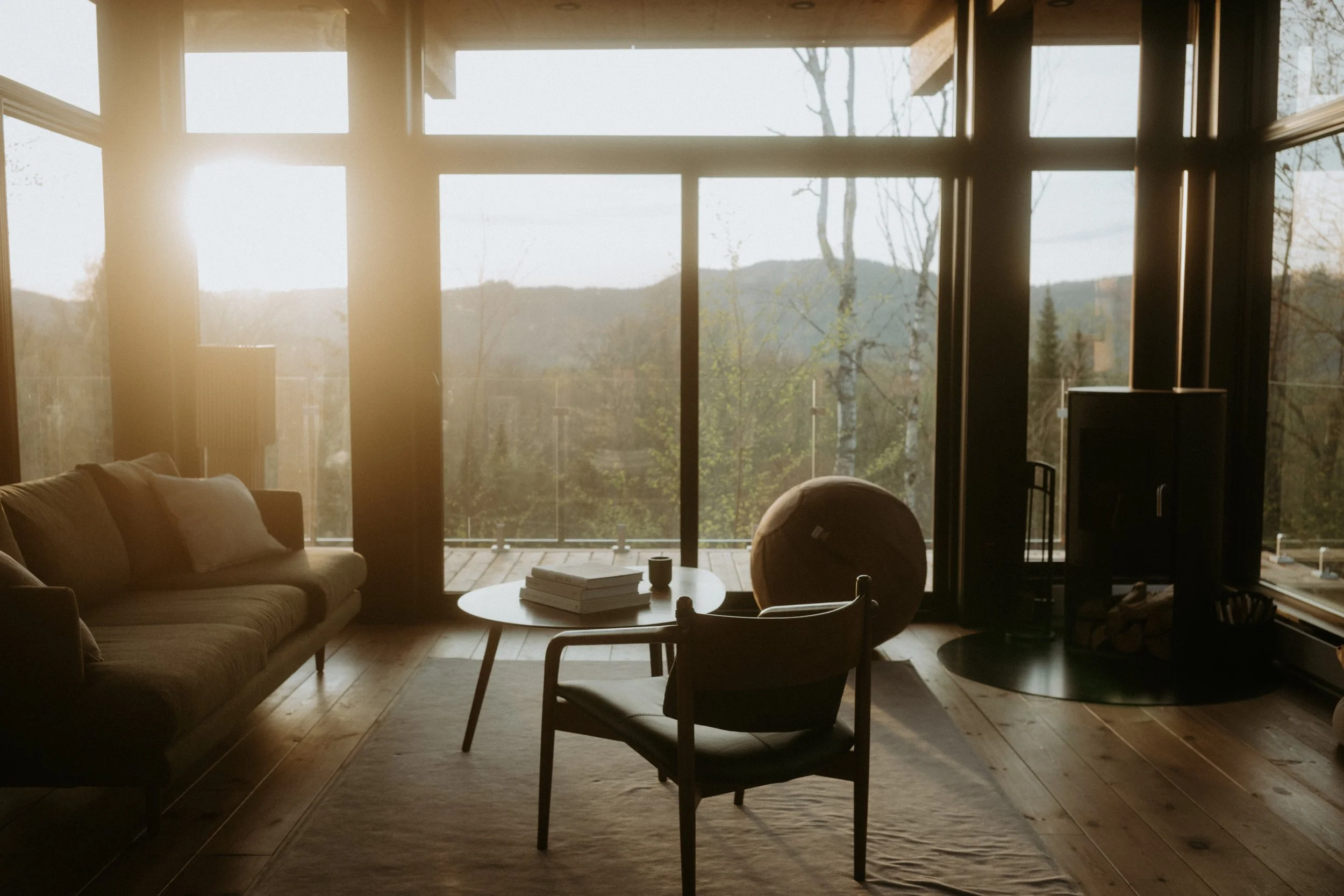 Living room with large windows letting in sunlight, a sofa on the left, a coffee table with books and a cup, a chair, and a round object in the corner, with a scenic view of trees and hills outside.