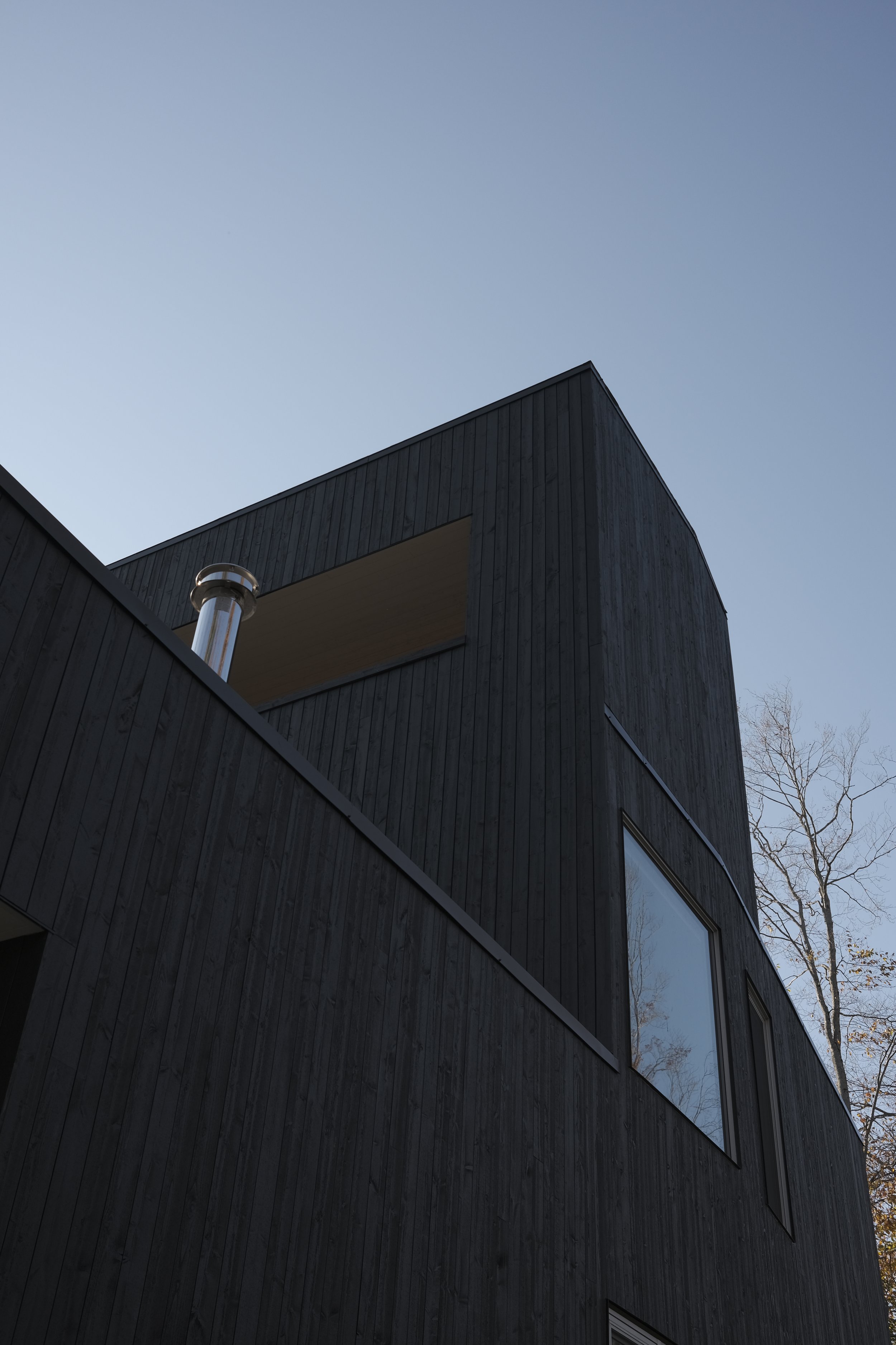 Close-up of a modern black wooden house with large windows and a chimney against a clear sky, with some trees visible in the background.