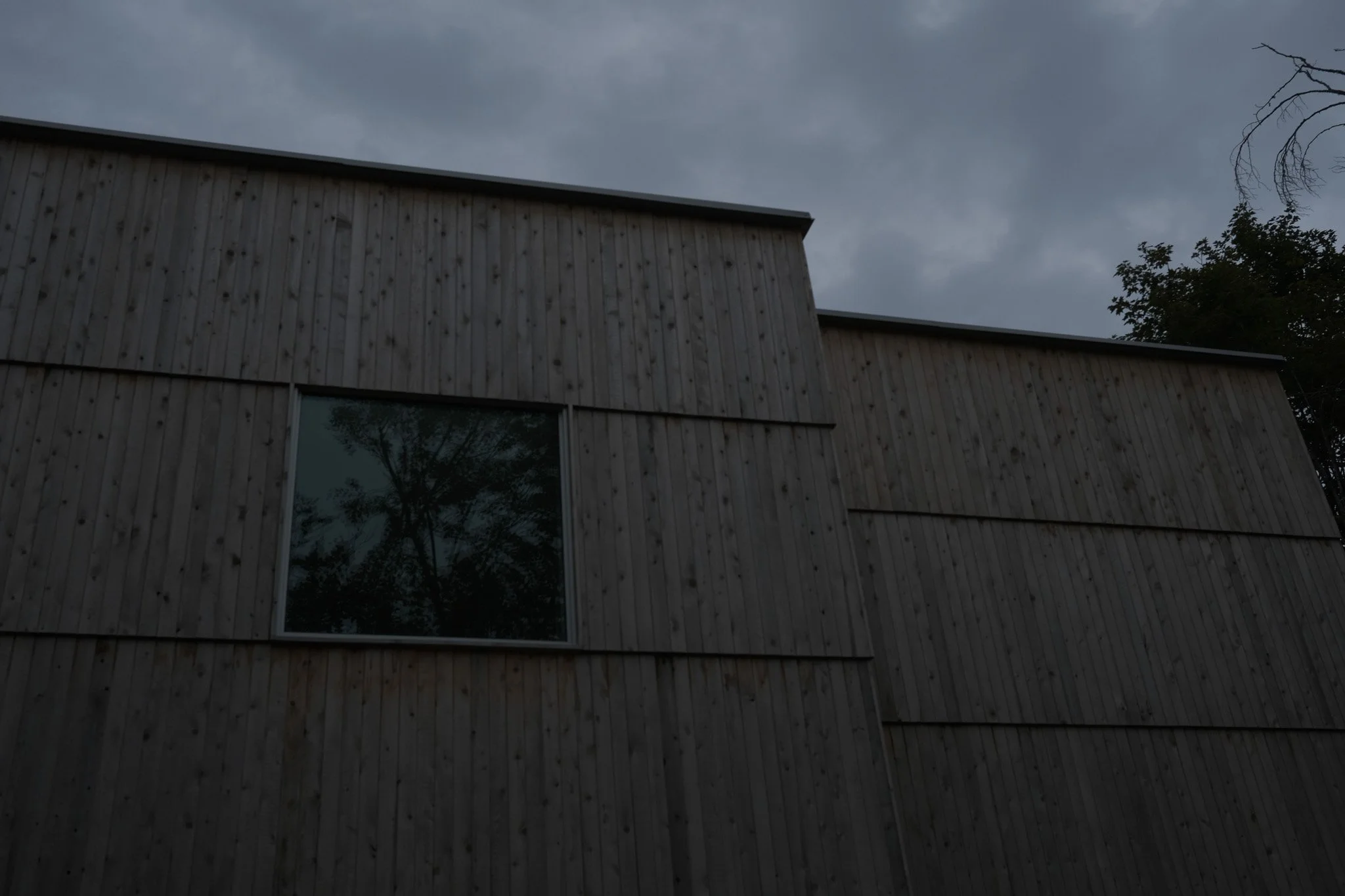 Modern building with wooden exterior and a window reflecting trees, under a cloudy sky.