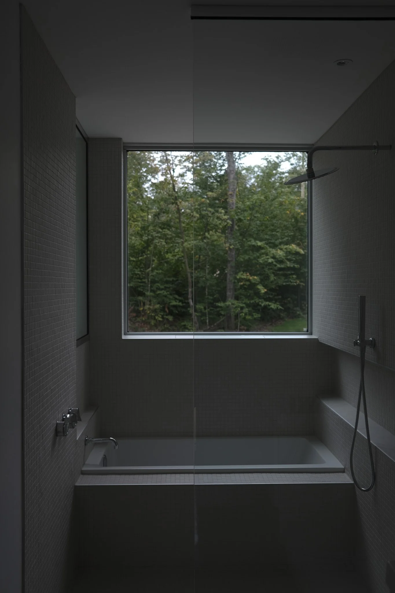 A modern bathroom with a bathtub beneath a large window overlooking a forest. The showerhead and faucet are on the right and left walls respectively, with gray mosaic tiles on the walls.