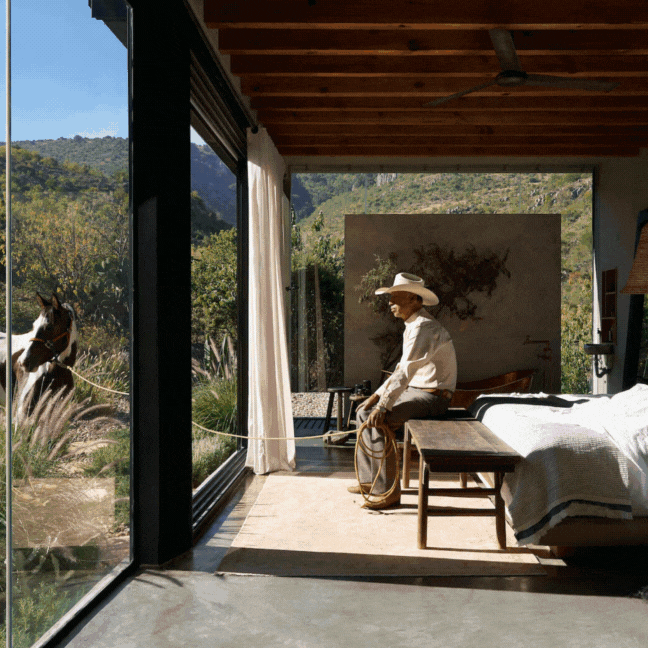 A person wearing a cowboy hat and white shirt sitting inside a modern room, looking out through glass doors at a horse outside in a natural landscape with hills and trees.