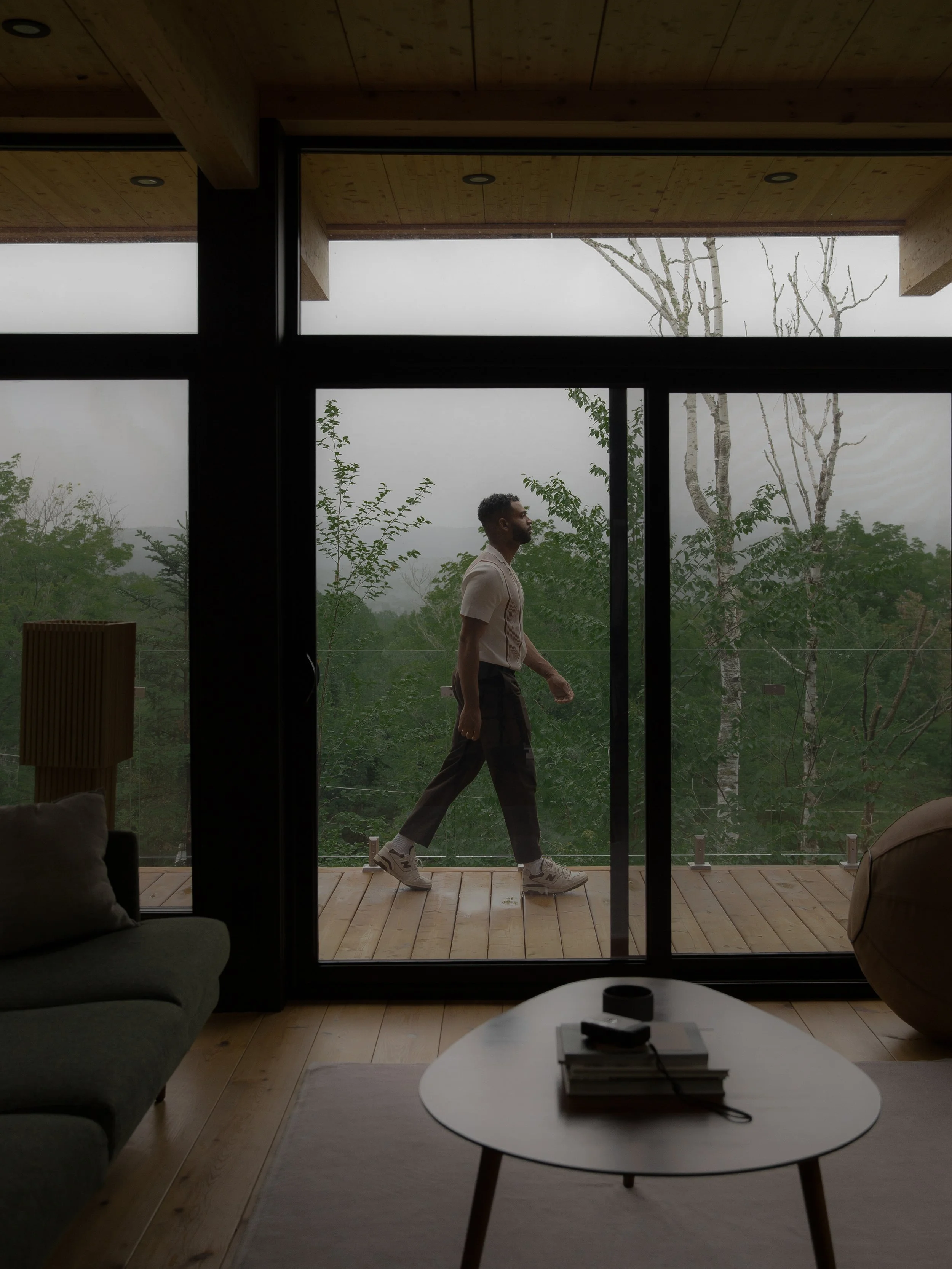 Man walking on wooden balcony outside a glass sliding door in a modern home with trees in the background.