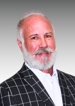 Portrait of an older man with white hair and a beard, wearing a black and white checkered blazer and a white shirt, smiling.