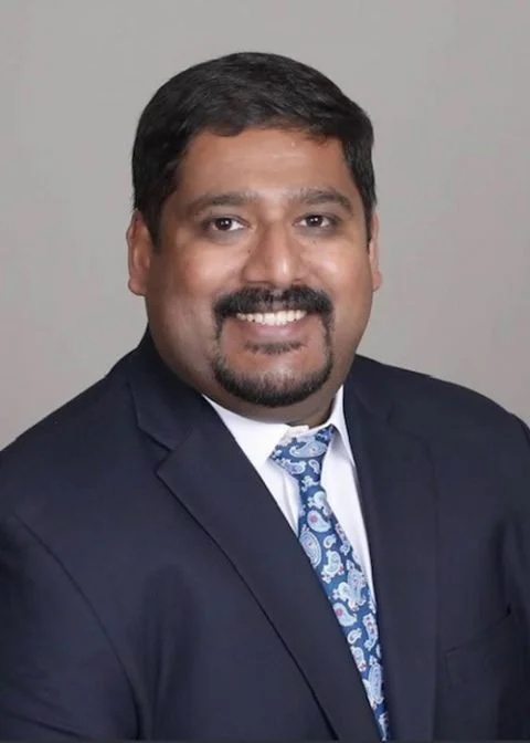 Professional man with short black hair and beard, wearing a dark suit, white shirt, and patterned tie, smiling against a plain gray background.