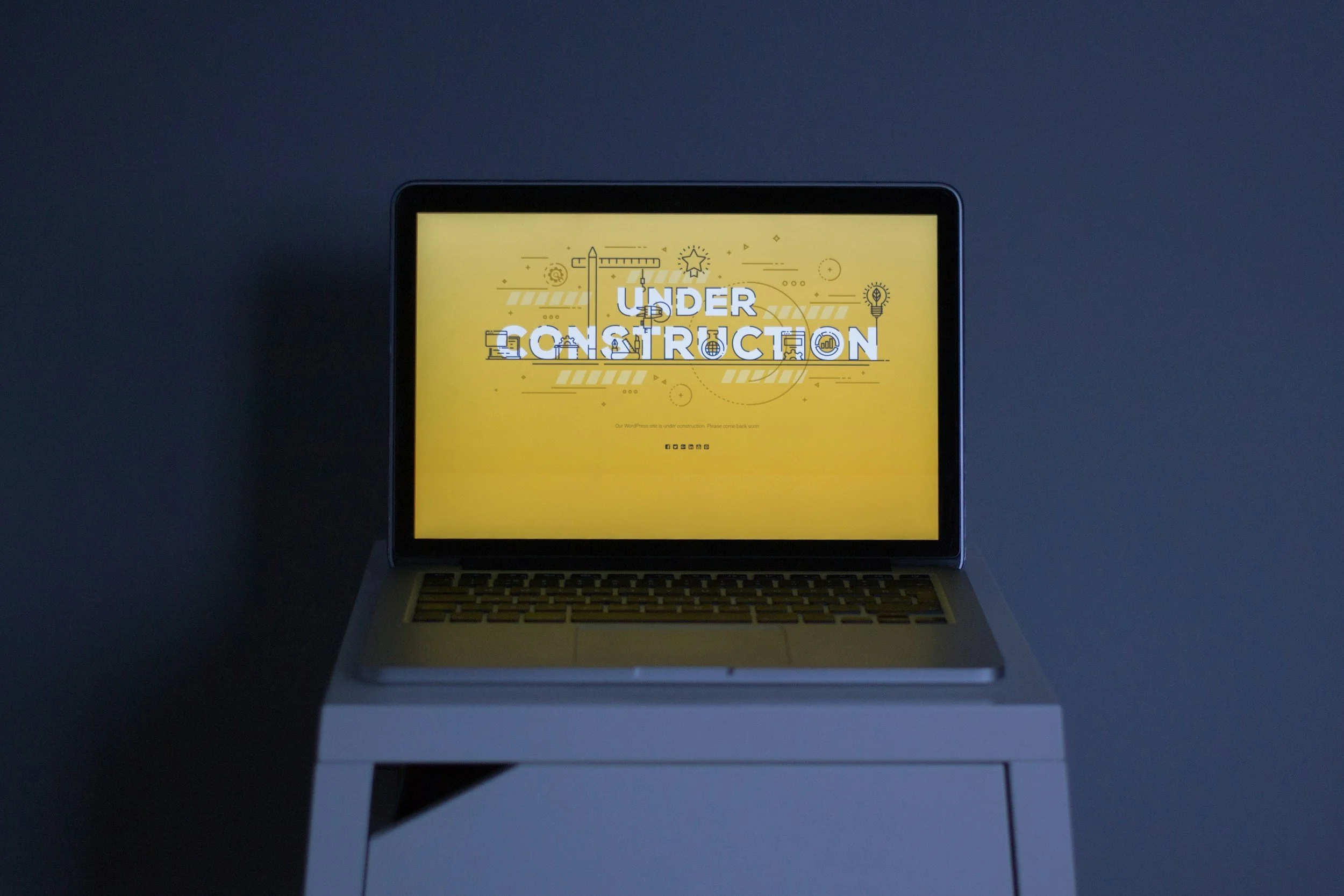 Laptop with a yellow screen displaying the text 'UNDER CONSTRUCTION' with construction-related icons, placed on a white desk in a dimly lit room.