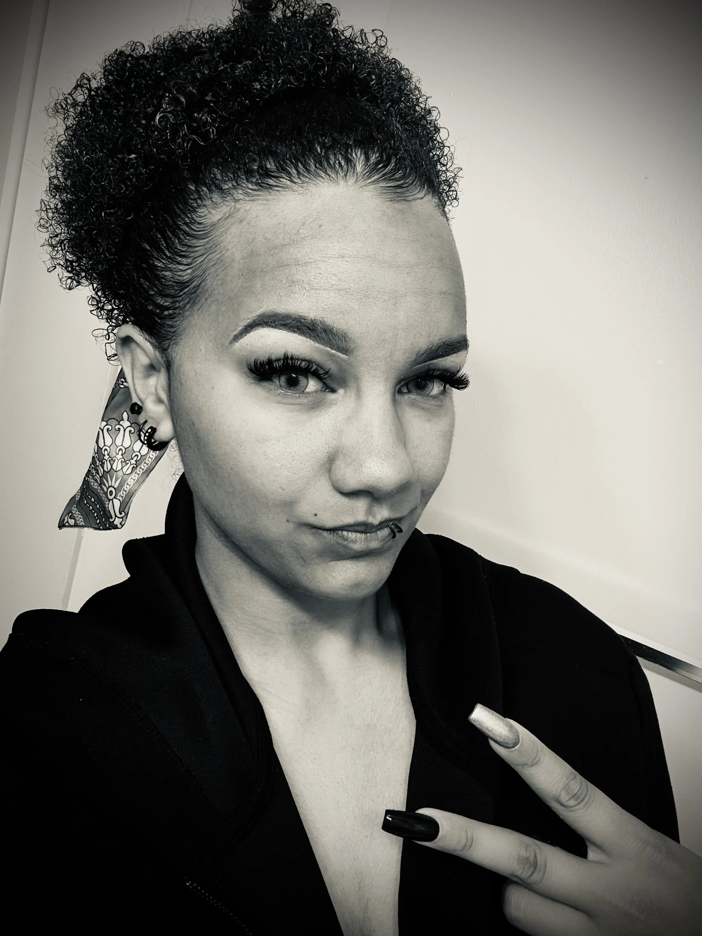 A woman with curly hair tied back with a bandana, makeup with prominent eyeliner, and long eyelashes, taking a selfie in black and white. She is holding up a peace sign with her fingers.
