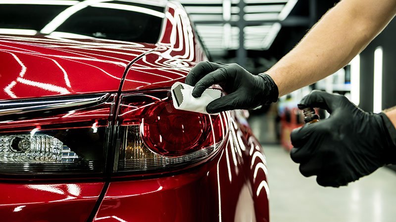 Person applying ceramic coating to a red car's taillight in a garage.