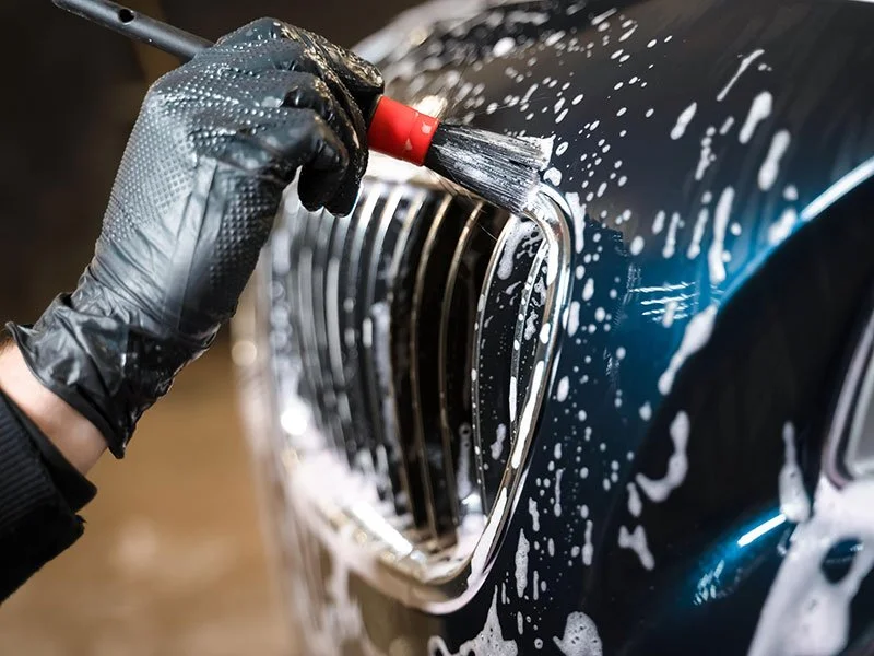 Close-up of a person cleaning a car's front grille with a brush and soap suds