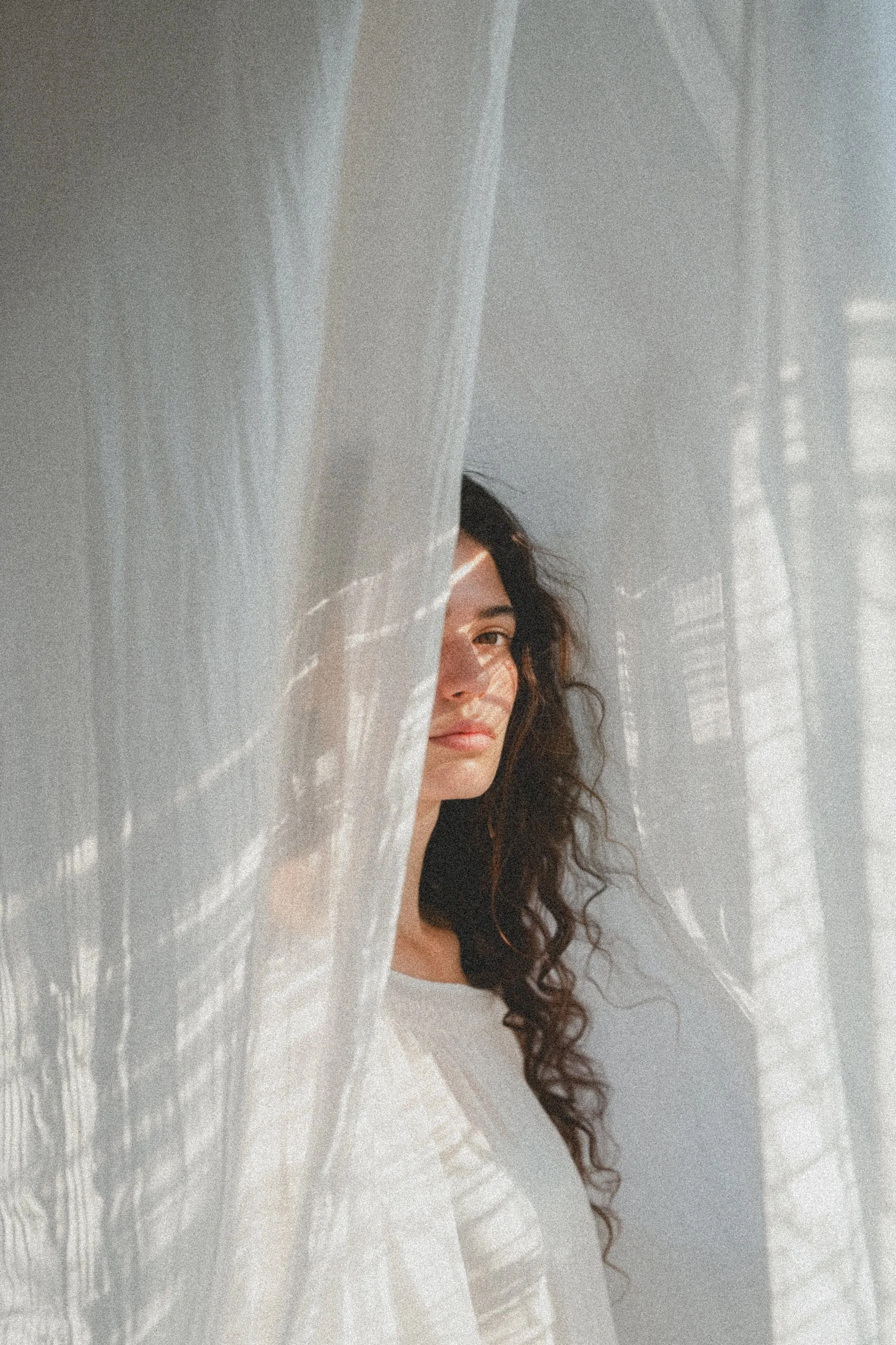 Woman with long curly hair standing behind sheer white curtains in soft natural light.