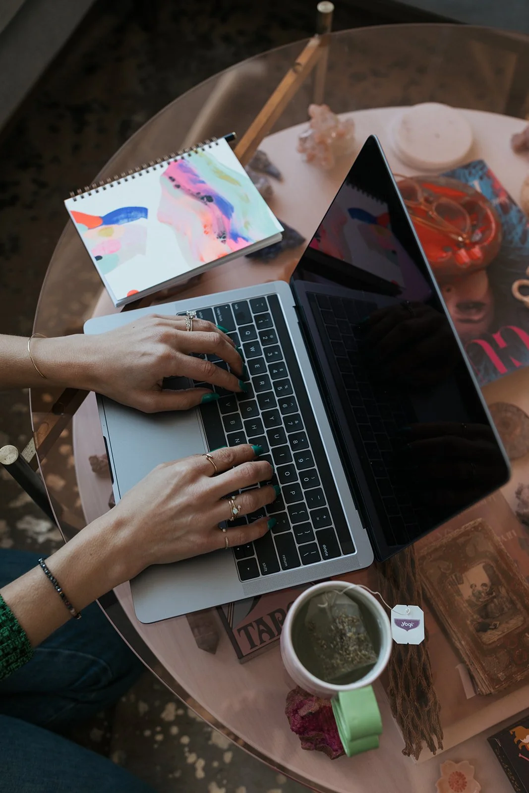 Person typing on a laptop at a glass table with a colorful notebook, a cup of tea, and scattered decorative items.