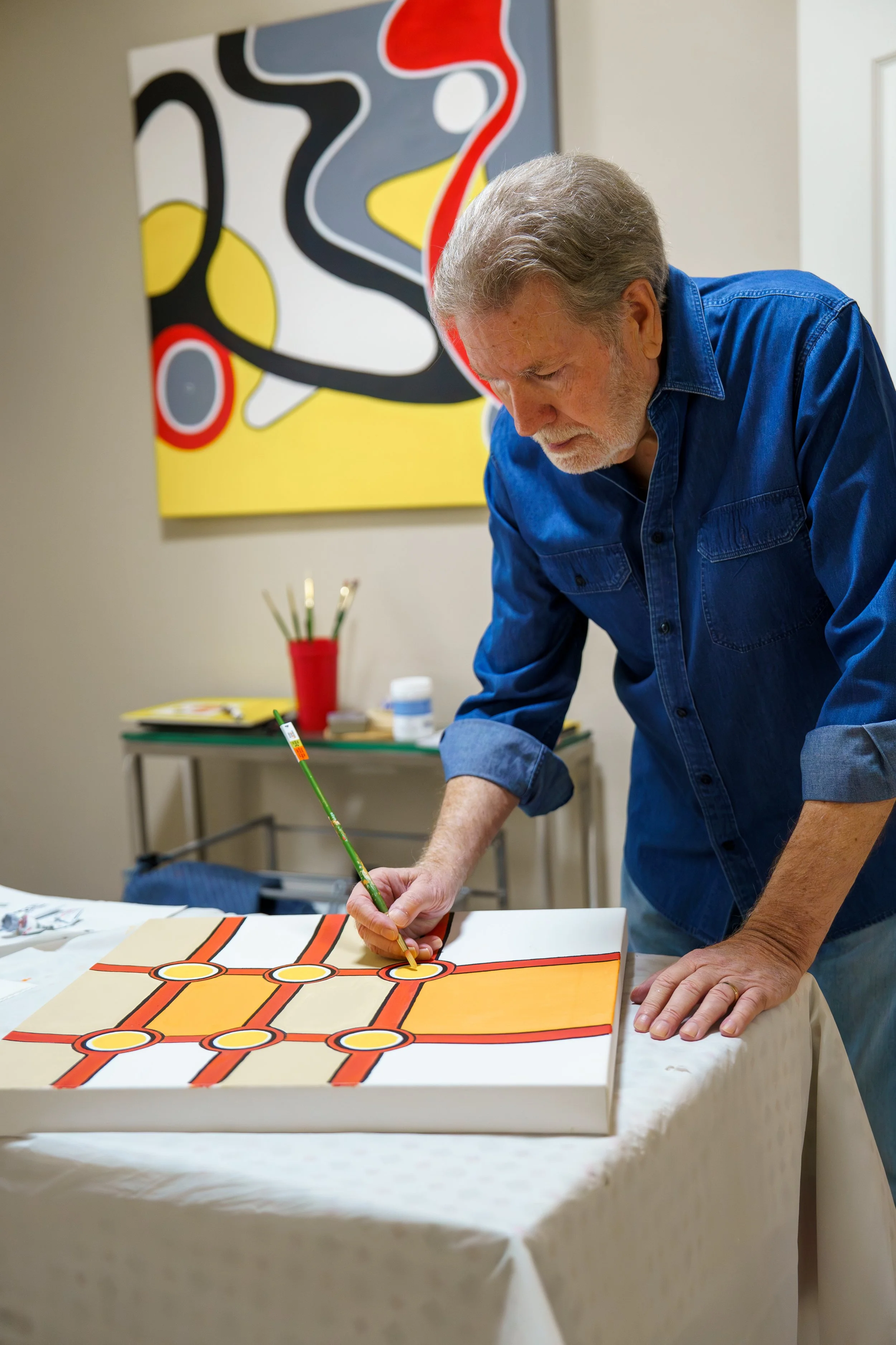 A man wearing a blue denim shirt is painting a colorful, geometric artwork on a canvas at a table in an art studio.