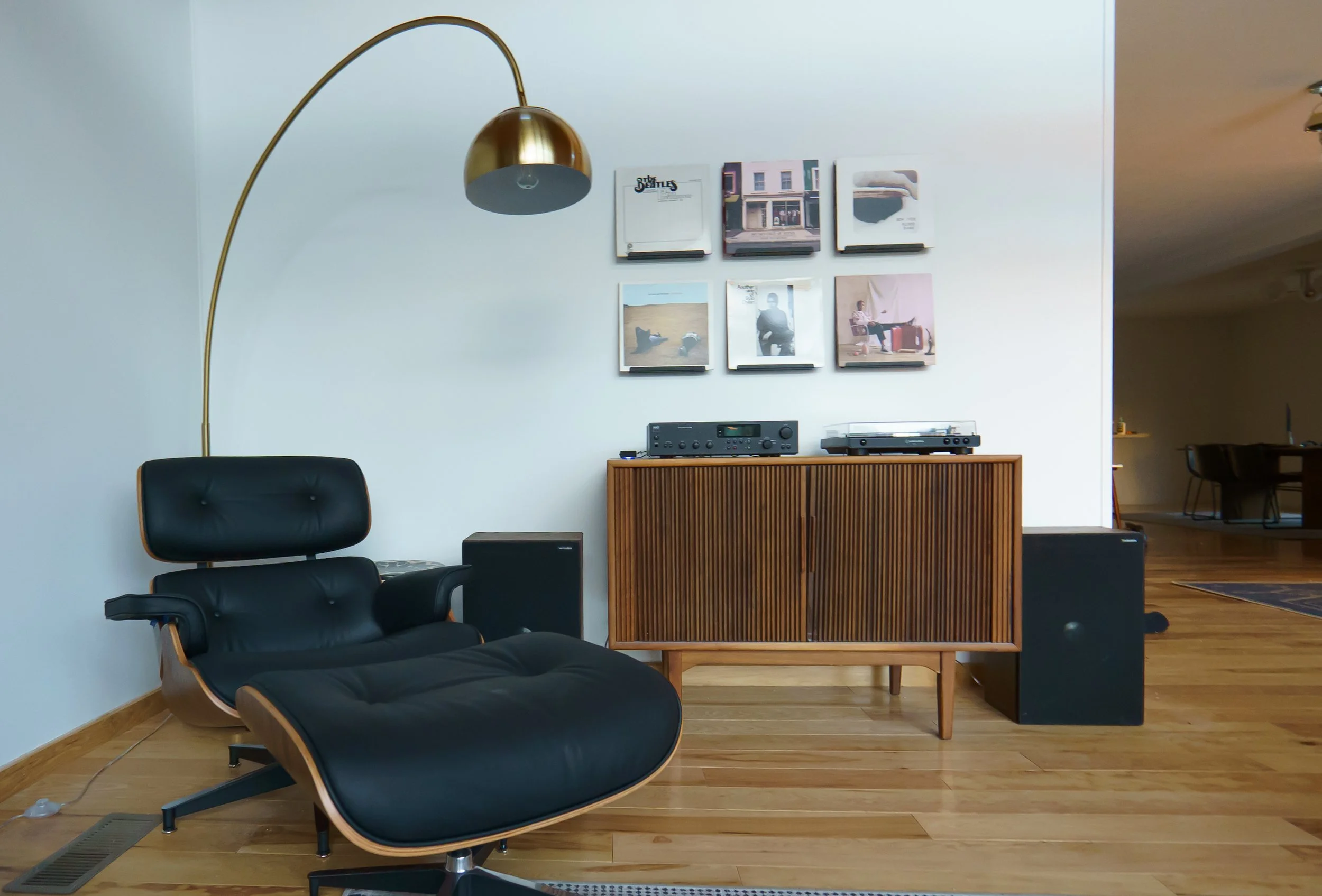 Eames lounge chair next to a wooden cabinet, topped by a stereo receiver and record player. Six shelves on the wall display records.