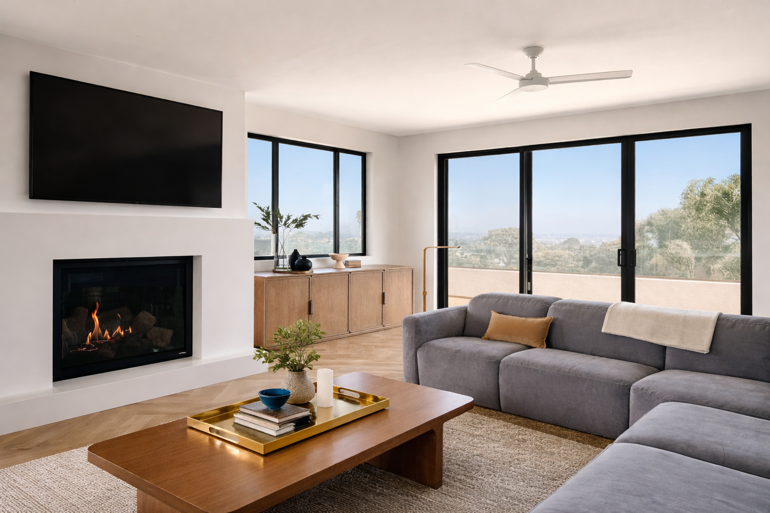Living room with a fireplace, large windows, and a sliding door leading to an outdoor balcony. There is a gray sectional sofa with a yellow pillow and cream blanket, a wooden coffee table with decorative items, and a wooden sideboard with vases and plants.