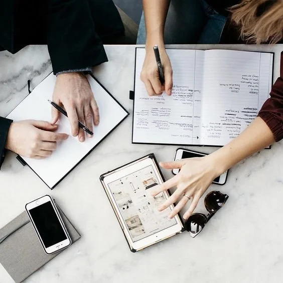 People working at a marble table with notebooks, a tablet, a smartphone, and sunglasses.