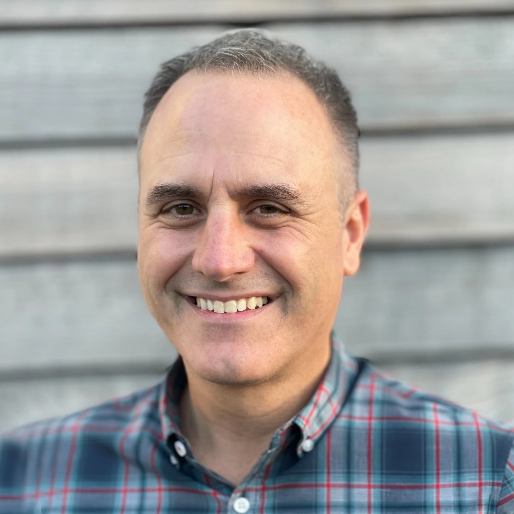 Portrait of a smiling man with short brown hair, wearing a plaid shirt, standing in front of a gray wooden wall.