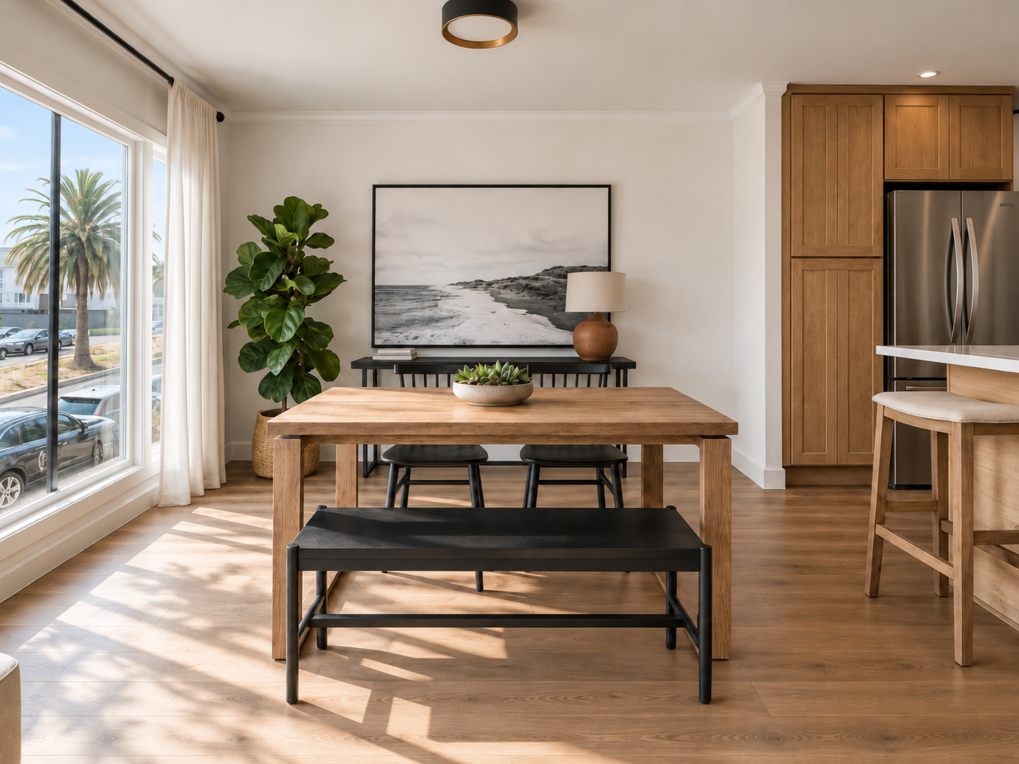 Bright dining room with large window, wooden flooring, wooden table with black bench, black chairs, green potted plant, black and white landscape art, side table with lamps and books, kitchen with wooden cabinets and stainless steel refrigerator.