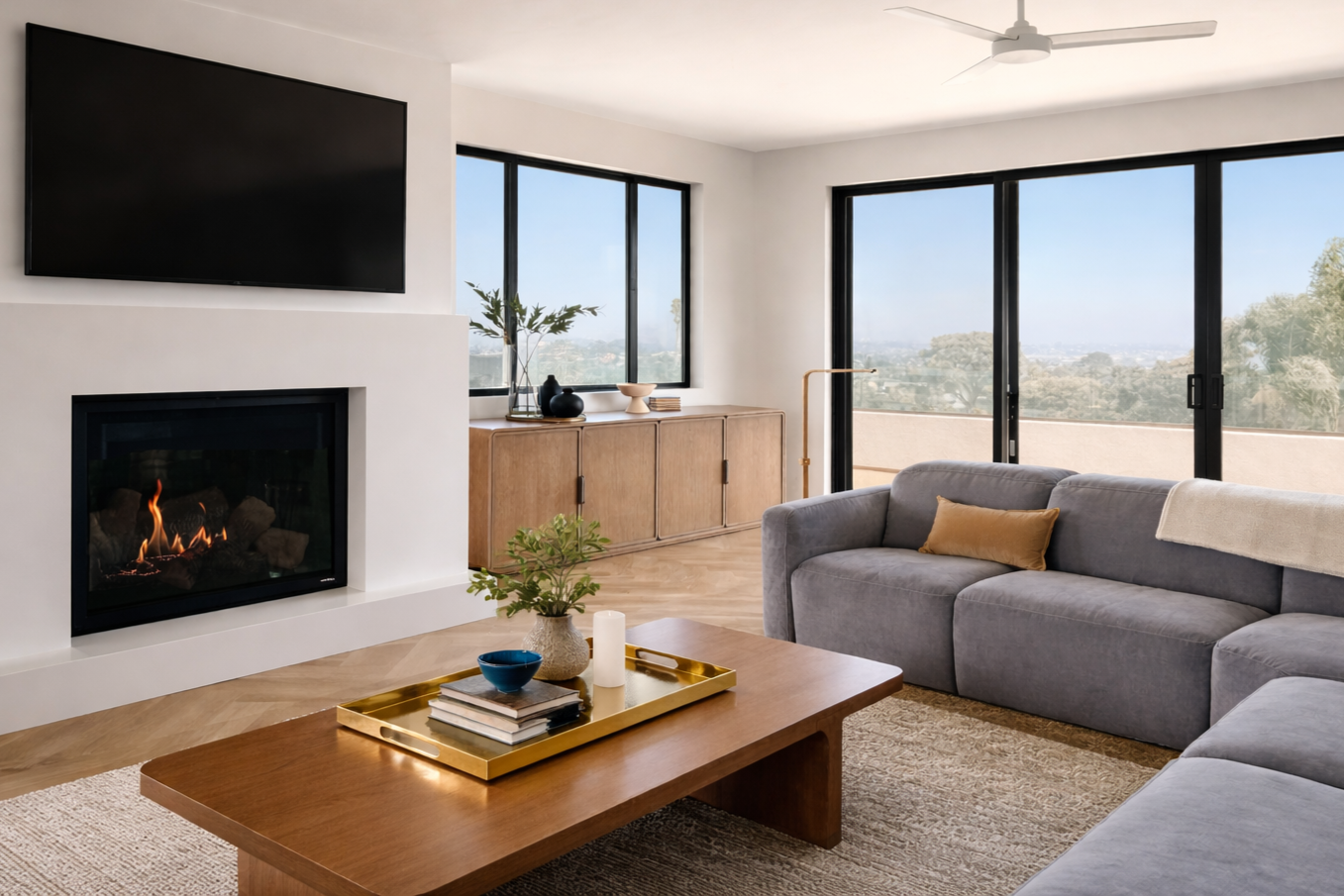 Living room with gray sectional sofa, wooden coffee table, fireplace, wall-mounted TV, large windows, sliding glass door, outdoor balcony with view, ceiling fan, decorative items on sideboard, indoor plants.